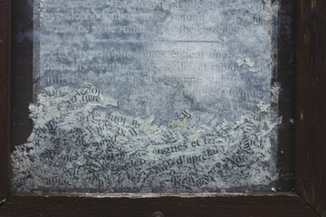 Close-up of weathered and partially erased text on a glass surface, framed by chipped dark wood. The faded black letters on a cloudy white background evoke age, decay, and forgotten information.