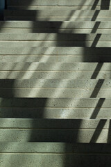 Concrete staircase lit by natural sunlight casting angular shadows through a railing. The image captures a minimalist architectural detail with strong contrast and linear patterns.