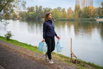 A determined woman carries empty plastic water bottles along a lakeside path, ready to fetch clean water. A scene depicting resilience and daily challenges.