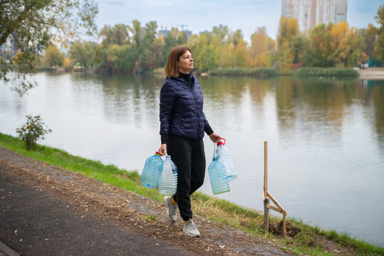 A determined woman carries empty plastic water bottles along a lakeside path, ready to fetch clean water. A scene depicting resilience and daily challenges.