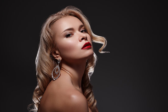 Elegant woman with wavy blonde hair, fashion red lipstick posing in flash lights against dark background. Diamonds jewelry, classic beauty, confidence and evening glamour. Hairstyle and makeup