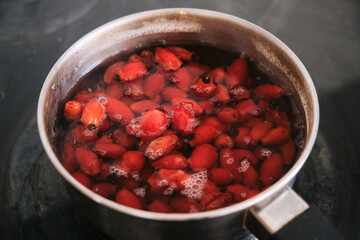 Close-up of red berries simmering in a metal pot on a stovetop, releasing vibrant juice and foam as they cook, suggesting homemade jam or fruit preserve preparation.