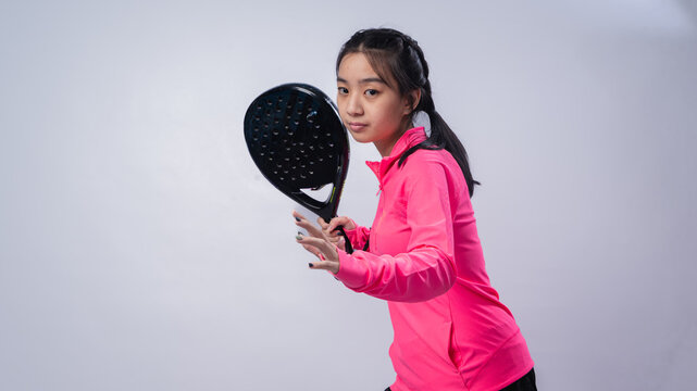 Asian woman in a pink long-sleeve sports outfit holding a black padel racket in a ready-to-play stance, looking focused and confident against a clean white studio background