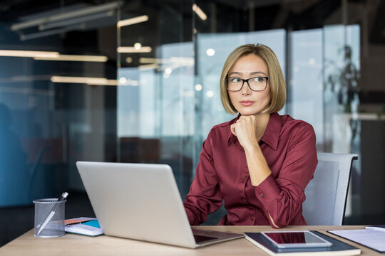Professional businesswoman in glasses sitting at her office desk, working on a laptop, and looking away thoughtfully, engaging in deep contemplation and strategic planning