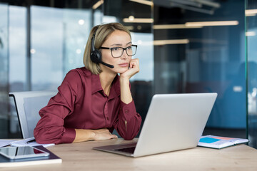 Bored woman with headset working on laptop, providing customer support while looking pensive, sitting at her desk in a modern corporate office environment