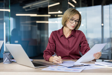 Businesswoman in a modern office desk, focused on analyzing financial reports and charts while writing notes, reviewing paperwork and planning strategy on a laptop