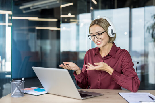 Woman with glasses and headphones smiling and gesturing during a laptop video call, working at a modern office desk in a focused, professional remote meeting setting
