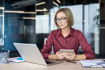 Businesswoman focused on work in a modern office, multitasking and interacting with technology devices while sitting at a wooden desk with a laptop and digital tablet