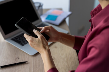 Woman's hands engaging with a smartphone while working at a desk, with a laptop open in the background, illustrating communication and business technology