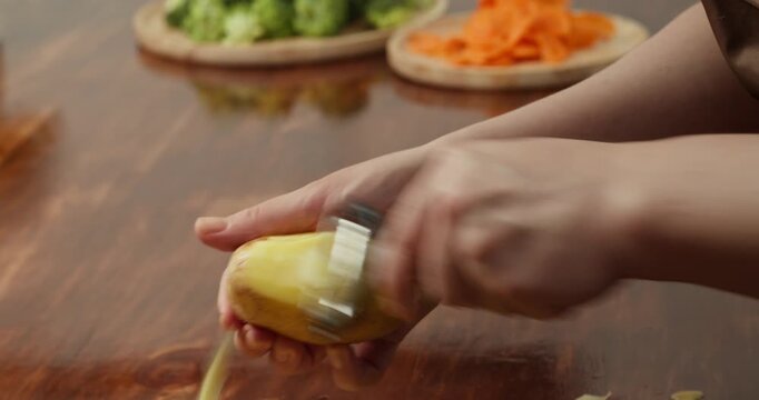 Hands peel a potato with a vegetable peeler over a wooden table. Concept of natural lifestyle, clean eating, and home cooking in a cozy rustic kitchen atmosphere.