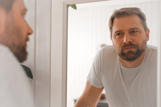 Confident handsome bearded man looking himself at the mirror. Pensive guy gazing thoughtfully. Captures introspection, grooming, hair care. Suitable for male health, lifestyle or personal care content