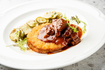 The dish consists of fried potato pancakes topped with a rich beef goulash and garnished with fresh greens and a side of sliced pickles or cucumbers.