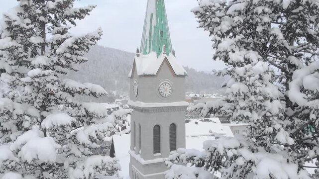 Aerial view of a snow-covered clock tower surrounded by tall evergreen trees, showcasing the serene winter landscape and gradual zoom out revealing the charming village below