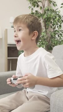 Seven year old boy sitting in armchair and playing video game with joystick, showing joy and excitement after winning