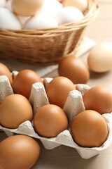 Many raw chicken eggs in carton and wicker basket on wooden table, closeup