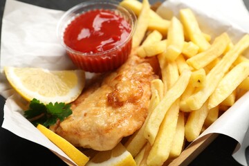 British Traditional Fish and chips with sauce, lemon and parsley in paper box on table, closeup