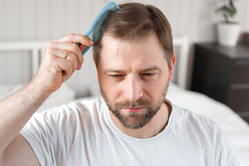 Obraz premium Handsome man combing brushing hair sitting at home light white bedroom. Guy styling grooming his hair with a comb. Self-care. Softly lit indoor space. Portrait. Male haircare morning routine concept