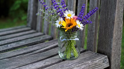 A colorful bouquet of flowers in a clear glass jar sits on a weathered wooden bench. Bright sunflowers, daisies, and lavender create a charming summer display in a lush garden - Powered by Adobe