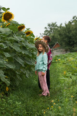 a girl in a straw hat and tracksuit stands near sunflowers and smiles
