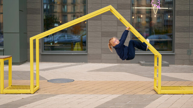 girl in blue hanging playfully on yellow metal structure outdoors. child enjoying playground fun, displaying youthful energy and joy. urban environment with modern design elements - Powered by Adobe