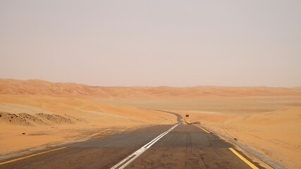Long empty highway stretching into the vast golden dunes of the Liwa Desert, United Arab Emirates. Scenic desert road disappearing into the horizon, surrounded by endless sand under a hazy sky. © nic
