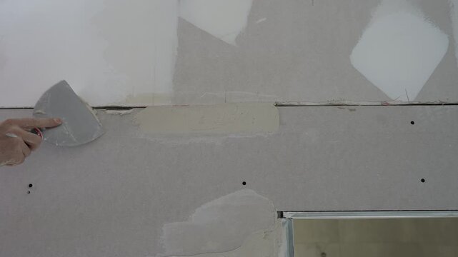 Close up of a worker using a metal spatula to apply plaster and fill joints between drywall panels during renovation in a new apartment. The process demonstrates accuracy, craftsmanship