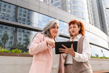 Fototapeta premium Two mature professional women reviewing data on tablet outside modern office building