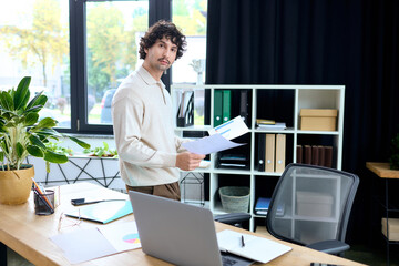Handsome young man standing in a modern office, holding documents and looking confident