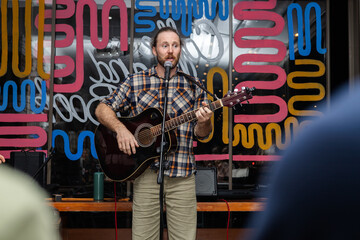 Fototapeta premium A young Caucasian man with a beard plays an acoustic guitar while singing on stage. Colorful abstract background enhances the lively atmosphere.