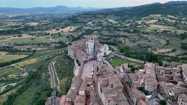 Aerial view of the Basilica of Saint Francis, a stunning architectural marvel nestled amidst rolling hills, contrasting with the tight-knit town, Assisi, Umbria, Italy.