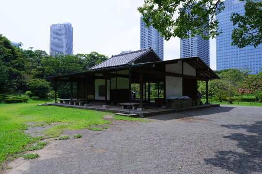A traditional wooden teahouse or rest shelter in a Japanese public garden, with modern high-rise buildings in the background