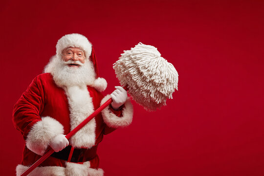 A cheerful Santa Claus in a red suit with a huge fluffy beard holds a large mop in his hands.