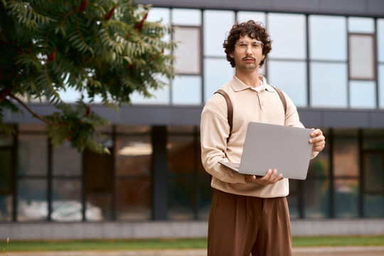 Young man in casual clothing stands with laptop outside a modern building on a sunny day - Powered by Adobe
