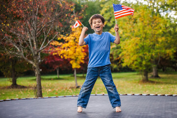 Smiling boy jumping high on trampoline outdoors holding Canada and USA flags in his hands on a bright autumn day