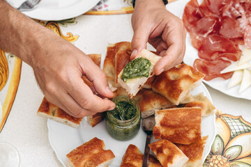 Hands of a man dipping bread into pesto sauce while enjoying a gourmet meal with assorted meats and cheeses on a beautifully decorated table