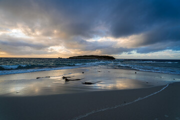 Reflets dorés sur le sable humide face à un ciel dramatique au-dessus de l'île, mer d'Iroise turquoise et nuages en mouvement sur une plage bretonne à marée basse.