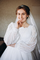 A beautiful bride in a white lace wedding dress smiles softly indoors. She stands by a window with sheer curtains, looking radiant and happy on her special day before the ceremony.