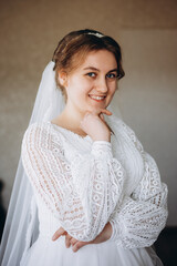 A beautiful bride in a white lace wedding dress smiles softly indoors. She stands by a window with sheer curtains, looking radiant and happy on her special day before the ceremony.
