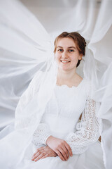 A smiling bride in a white lace wedding dress poses with her veil flowing gracefully around her. The soft fabric and natural light create a romantic and dreamy bridal portrait.