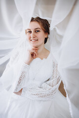 A smiling bride in a white lace wedding dress poses with her veil flowing gracefully around her. The soft fabric and natural light create a romantic and dreamy bridal portrait.