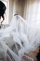 Bride in a luxurious white dress stands by the window, her veil gently flowing in the air. A scene filled with tenderness, light, and anticipation of a special wedding moment.