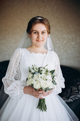 A beautiful bride in a white lace wedding dress holds a bouquet of white roses and greenery. She smiles softly, posing indoors before the wedding ceremony, looking elegant and serene.