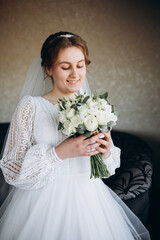 A beautiful bride in a white lace wedding dress holds a bouquet of white roses and greenery. She smiles softly, posing indoors before the wedding ceremony, looking elegant and serene.
