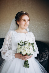 A beautiful bride in a white lace wedding dress holds a bouquet of white roses and greenery. She smiles softly, posing indoors before the wedding ceremony, looking elegant and serene.