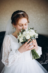A beautiful bride in a white lace wedding dress holds a bouquet of white roses and greenery. She smiles softly, posing indoors before the wedding ceremony, looking elegant and serene.