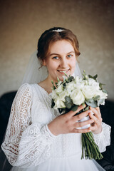 A beautiful bride in a white lace wedding dress holds a bouquet of white roses and greenery. She smiles softly, posing indoors before the wedding ceremony, looking elegant and serene.