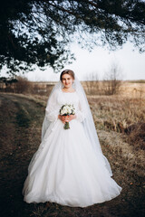 A bride in a beautiful white lace dress stands outdoors holding a bouquet of white roses. A calm and tender atmosphere of a wedding day in nature.