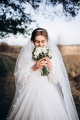A bride in a beautiful white lace dress stands outdoors holding a bouquet of white roses. A calm and tender atmosphere of a wedding day in nature.