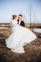 The groom lifts the bride in his arms outdoors as they smile at each other. Her flowing white dress creates a romantic feel, capturing a warm and joyful moment from their wedding photoshoo