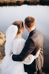 The newlyweds stand by the water, gently embracing as they look into the distance. The bride in a lace dress and veil and the groom in a dark suit create a serene and romantic outdoor moment.